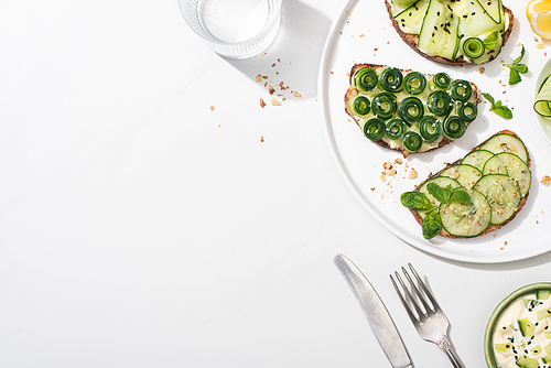 top view of fresh cucumber toasts with seeds, mint and basil leaves on plate near cutlery, water and yogurt on white background