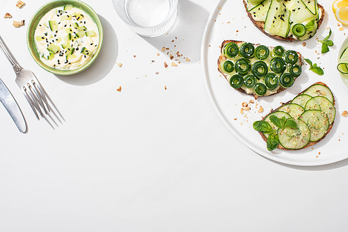 top view of fresh cucumber toasts on plate near lemon, water, cutlery and yogurt on white background