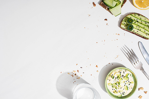 top view of fresh cucumber toasts near lemon, water, cutlery and yogurt on white background