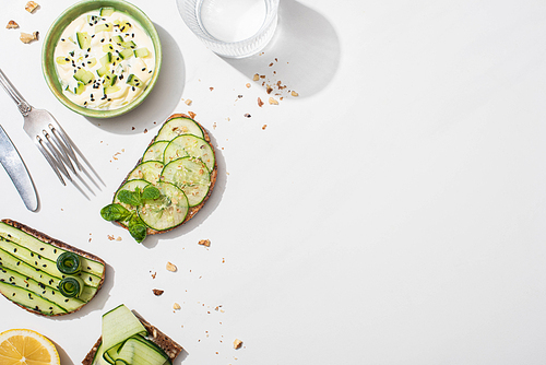 top view of fresh cucumber toasts near lemon, water, cutlery and yogurt on white background