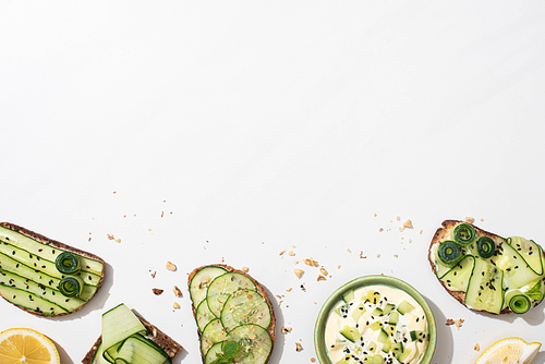 top view of fresh cucumber toasts with seeds, mint and basil leaves and lemon near bowl of yogurt on white background