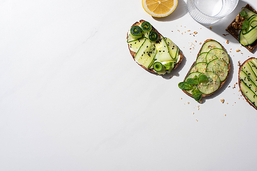 top view of fresh cucumber toasts with seeds, mint and basil leaves, lemon near water on white background