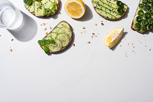 top view of fresh cucumber toasts with seeds, mint and basil leaves, lemon near water on white background