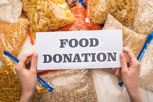 cropped view of woman holding card with food donation lettering near groats and pasta in zipper bags