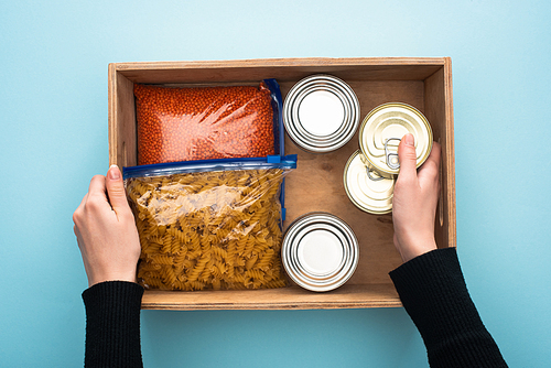 cropped view of woman holding wooden box with cans and groats in zipper bags on blue background