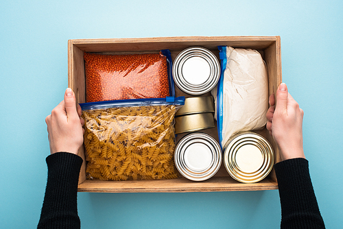 cropped view of woman holding wooden box with cans and groats in zipper bags on blue background
