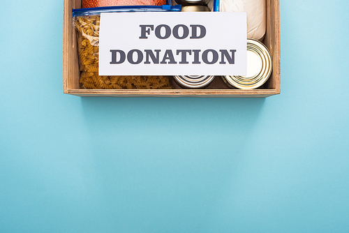 top view of cans and groats in zipper bags in wooden box near card with food donation lettering on blue background