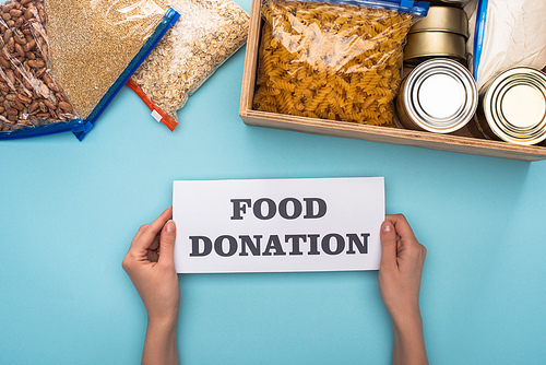 cropped view of woman holding card with food donation lettering near cans and groats in zipper bags in box on blue background