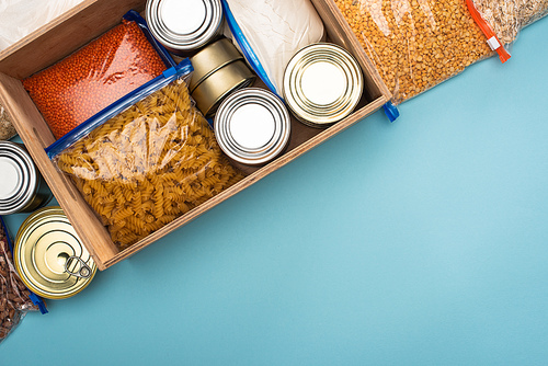 top view of cans and groats in zipper bags in wooden box on blue background with copy space, food donation concept