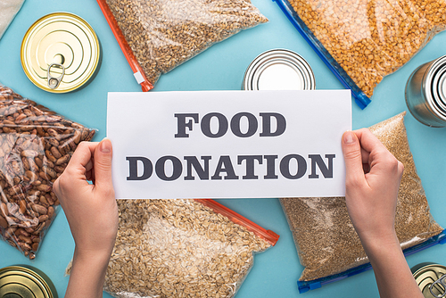 cropped view of woman holding card with food donation lettering near cans and groats in zipper bags on blue background