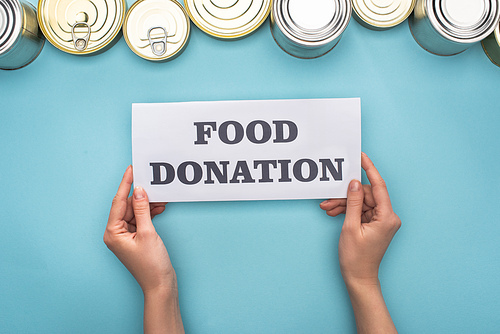 cropped view of woman holding card with food donation lettering near cans on blue background