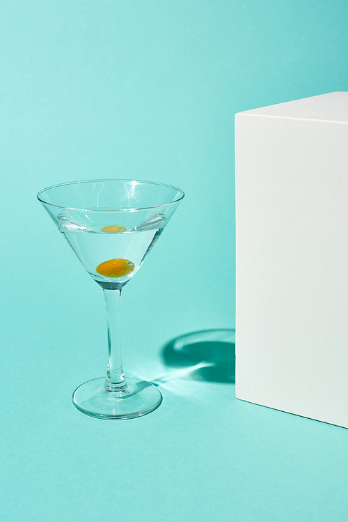 transparent glass with cocktail and olive near white cube on turquoise background