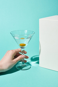 cropped view of woman holding transparent glass with cocktail and olive near white cube on turquoise background