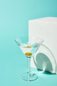 transparent glass with cocktail and olive near white cube on turquoise background