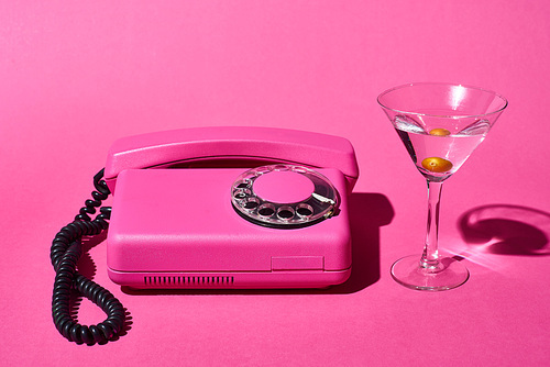 transparent glass with cocktail and olive near vintage dial phone on pink background