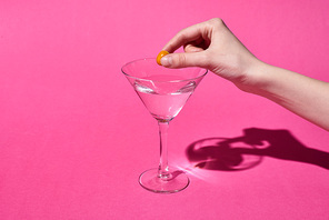 cropped view of woman putting olive in glass with cocktail on pink background