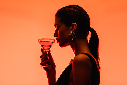 side view of young woman holding margarita cocktail with ice cubes on orange