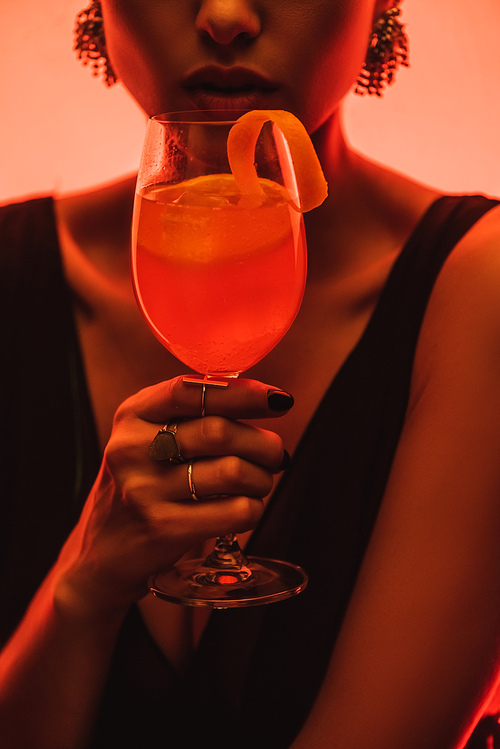 partial view of elegant woman holding glass with mixed cocktail and orange on pink