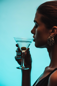 side view of woman holding glass and drinking martini isolated on blue