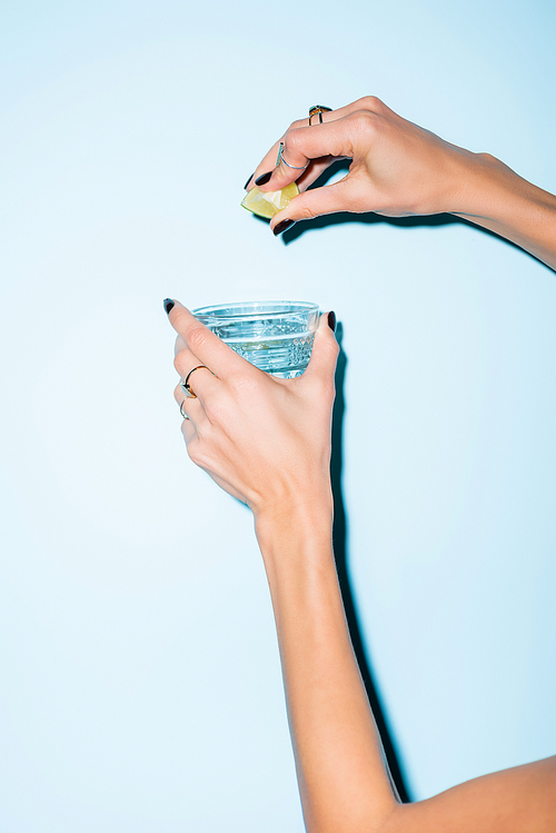 cropped view of woman squeezing lime in glass with alcohol drink on blue