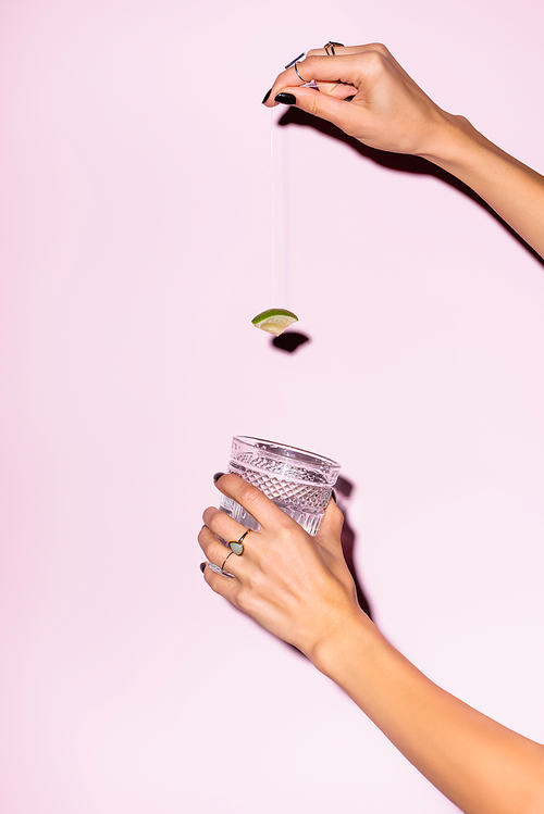cropped view of woman holding lime above glass with alcohol drink on pink