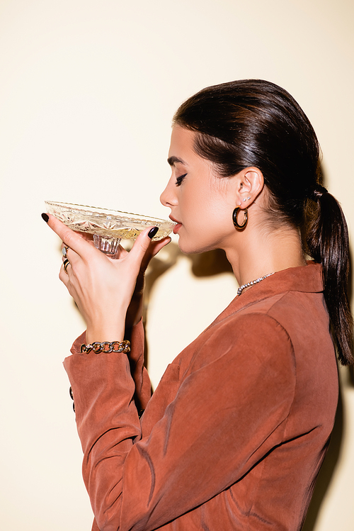side view of brunette woman in brown blazer drinking champagne from glass on white