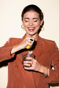 happy woman in brown blazer squeezing lemon in glass with alcohol cocktail on white