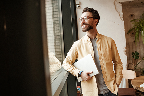 bearded freelancer smiling and holding laptop while standing with hand in pocket near window