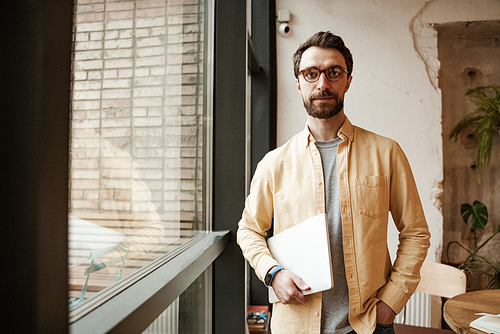 bearded freelancer holding laptop while standing with hand in pocket near window