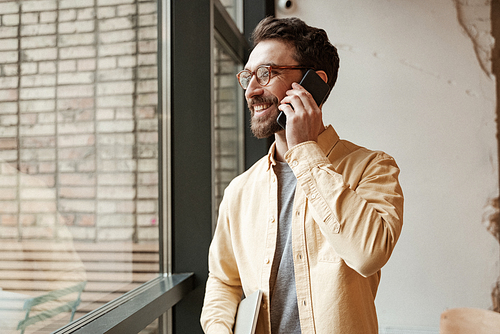 happy and bearded freelancer holding laptop while standing near window and talking on smartphone