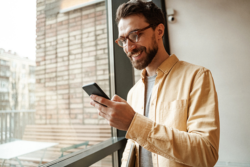 smiling and bearded man using smartphone while standing near window