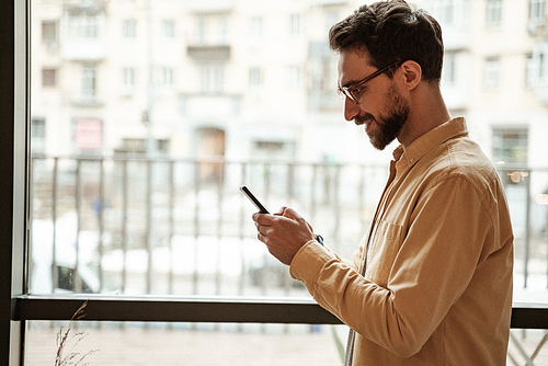 side view of bearded man using smartphone while standing near window