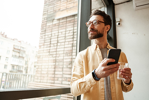 bearded man holding smartphone while standing near window with glass of water
