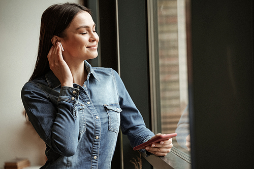 pleased woman in wireless earphones listening music and holding smartphone