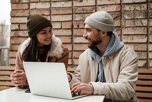 cheerful freelancer in beanie hats looking at each other near laptop