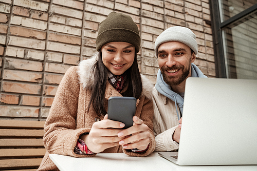 happy freelancers in beanie hats looking at smartphone near laptop on table