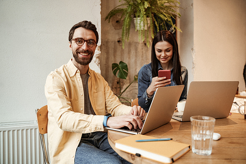 happy freelancer in eyeglasses using laptop near woman in cafe