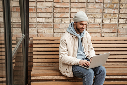 happy bearded freelancer in beanie hat using laptop while sitting on bench