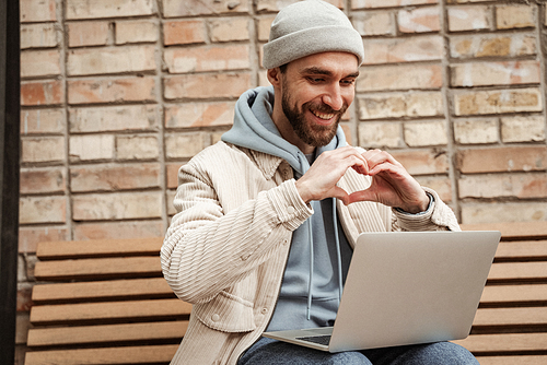 happy bearded freelancer in beanie hat showing heart sign while using laptop and sitting on bench