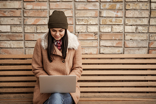 happy brunette freelancer in beanie hat using laptop while sitting on bench