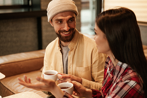 bearded man in beanie hat holding cup of coffee and taking with woman on blurred foreground