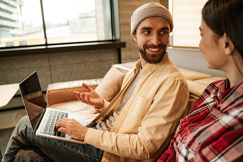 happy bearded freelancer in beanie hat gesturing near laptop and woman
