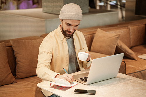 bearded freelancer holding cup of coffee and writing in notebook near laptop