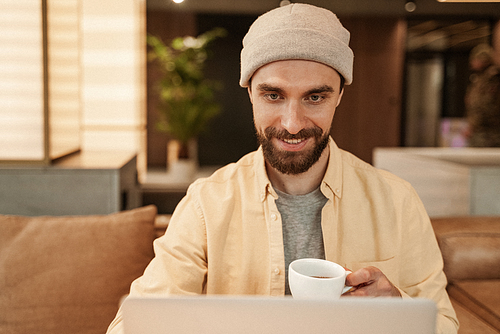bearded freelancer holding cup of coffee and using laptop