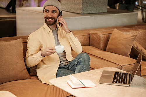 cheerful bearded man in beanie hat holding cup and talking on smartphone