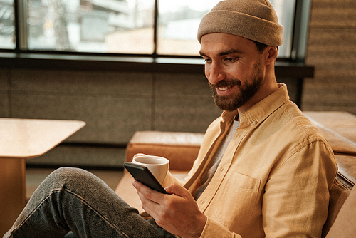 cheerful bearded man in beanie hat holding cup and using smartphone