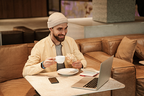 happy freelancer holding croissant and cup near gadgets on table