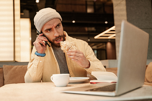 bearded freelancer eating croissant while talking on smartphone near laptop
