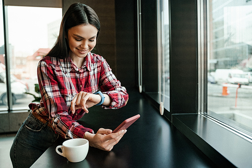 happy woman looking at smart watch while holding mobile phone near cup on table