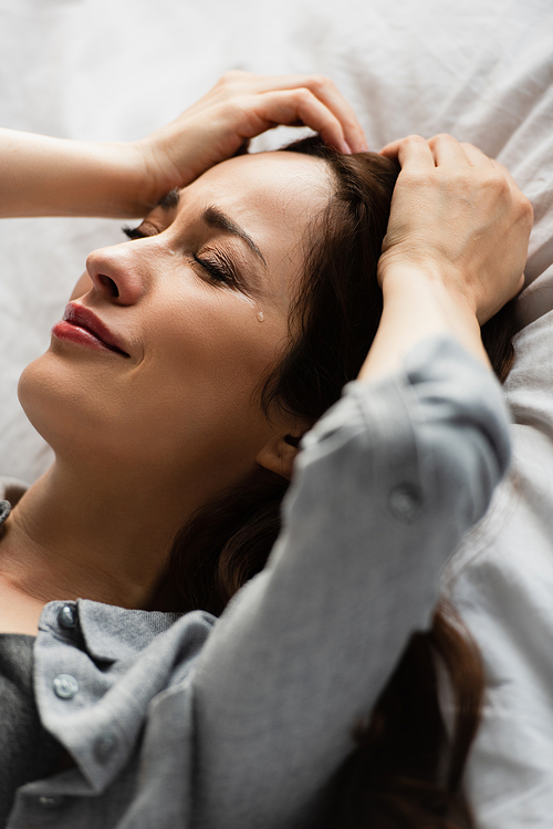 frustrated and brunette woman with closed eyes touching hair while crying on bed